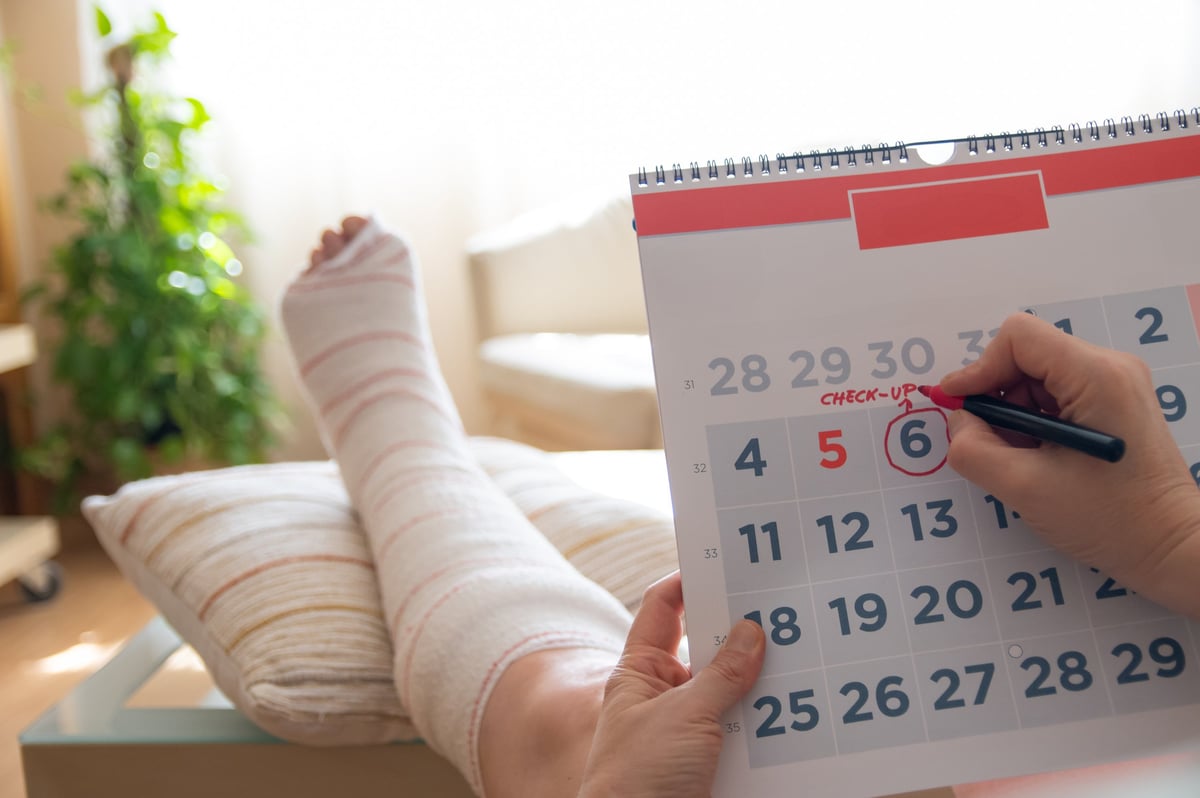 Patient with a broken leg rests their foot on a pillow, marking a date on a calendar for a medical check-up appointment, highlighting the importance of timely healthcare