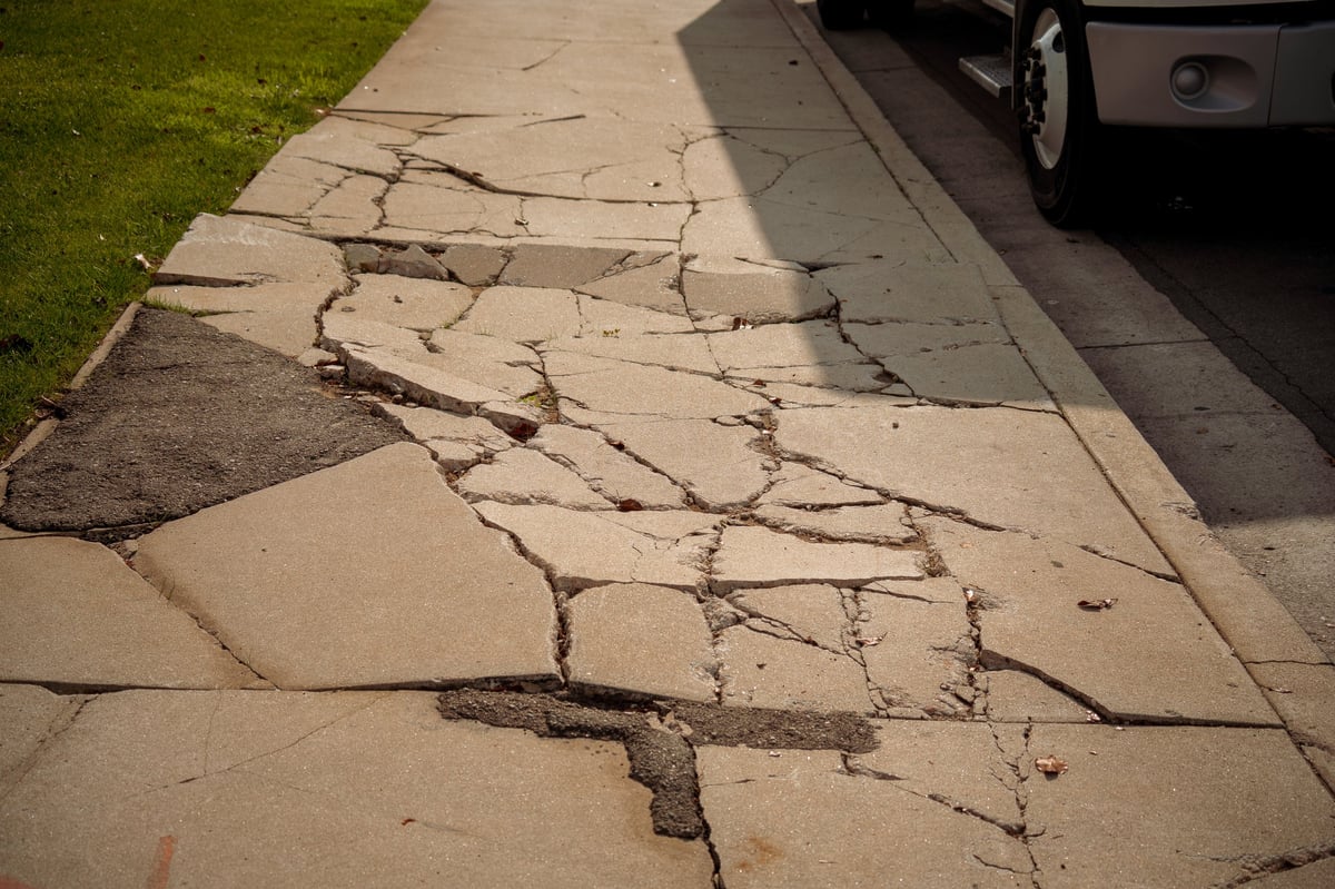 A damaged concrete sidewalk with large cracks and broken pieces, adjacent to a grassy area and a street with a vehicle tire